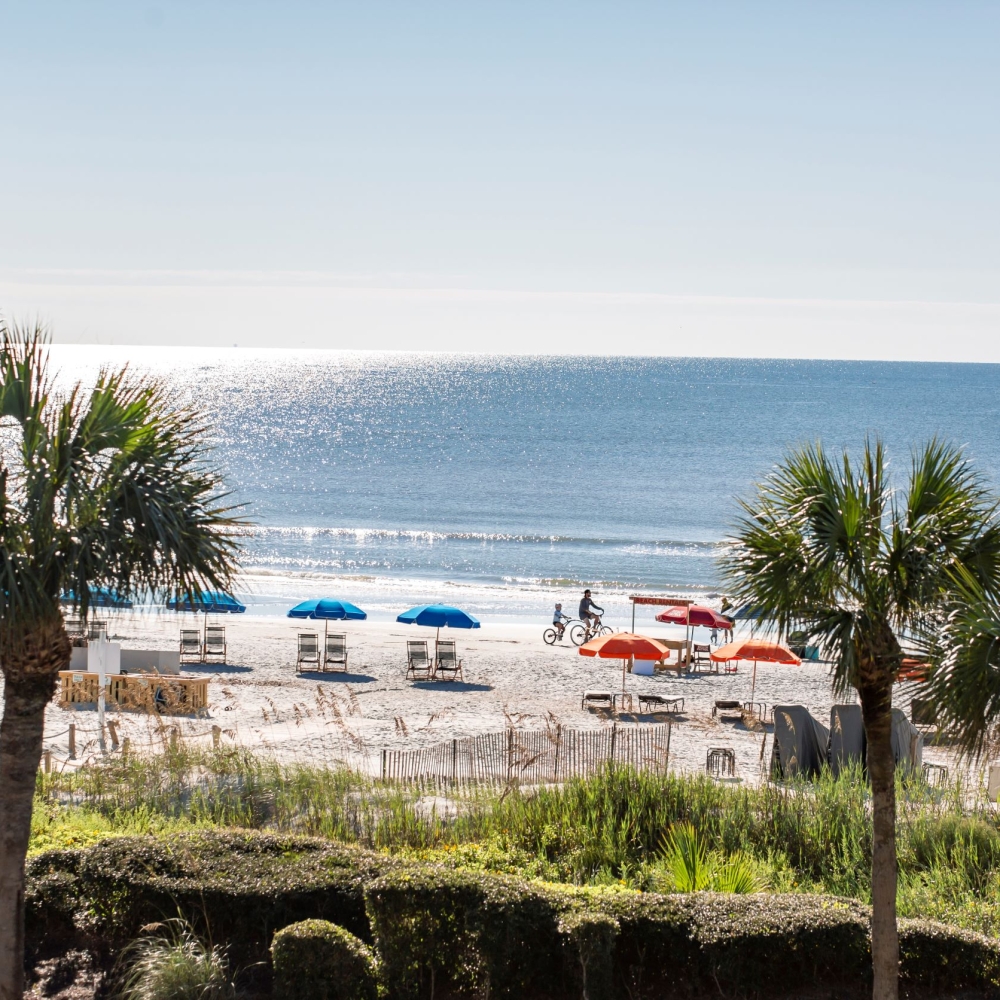 The view from the Sea Pines Beach Club of the beach with blue and orange umbrellas and a lifeguard stand and the ocean. 